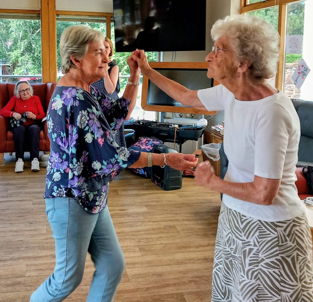 Sandra Currie and a friend dance in the lounge on a bright afternoon, both smiling as other women observe from the background. Sunlight pours through large windows onto the wooden flooring.