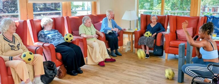 Six elderly women sit in armchairs doing movement exercises with yellow footballs, guided by a facilitator in a bright room with large windows letting in Jamaican sunshine.