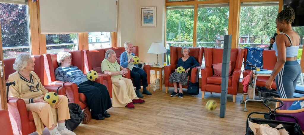 A group of elderly people sit in chairs holding yellow footballs as a woman encourages movement, leading a lively seated activity in a bright room flooded with Jamaican sunshine through large windows.