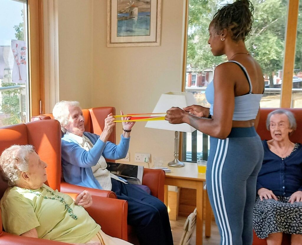 A woman in sportswear helps an elderly lady with a resistance band exercise at Lear House, bathed in Jamaican sunshine, whilst two other elderly women sit nearby, enjoying the activity.