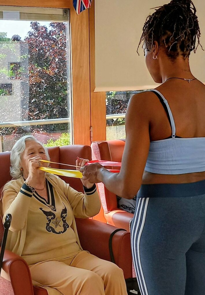 An elderly woman in an armchair holds one end of a resistance band as a younger woman in sportswear encourages her through a seated exercise, making new memories together indoors.