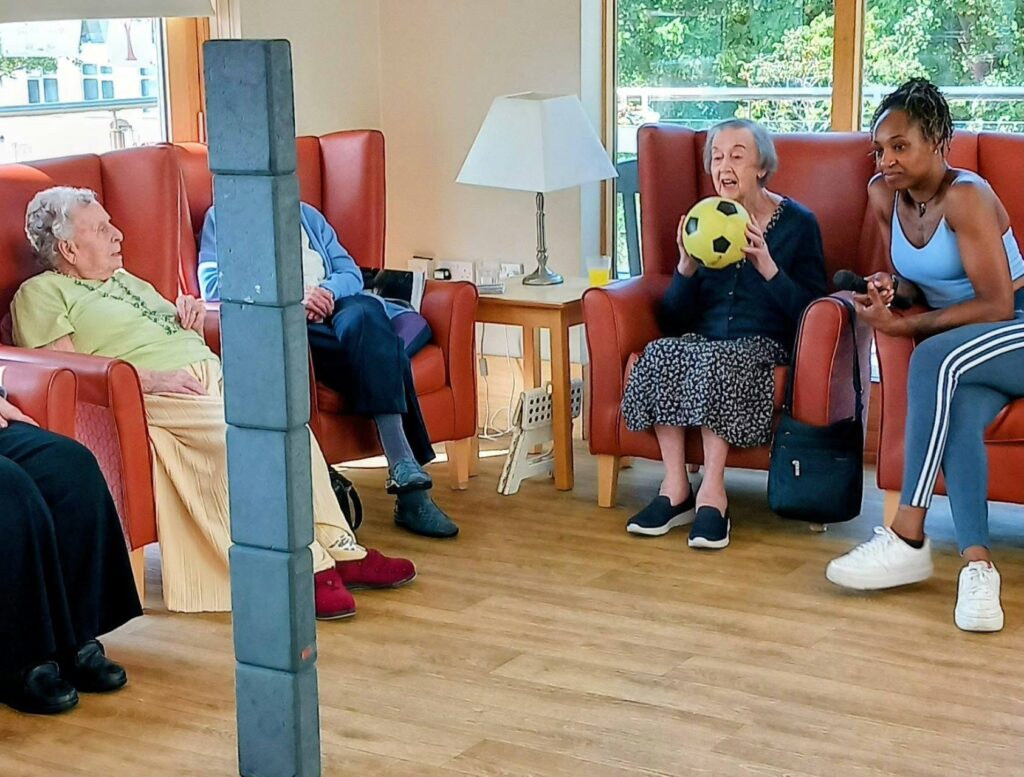 Several elderly women and a younger woman sit in armchairs indoors, reminiscing as one older lady holds a yellow and black football, with a stack of soft blocks visible in the foreground.