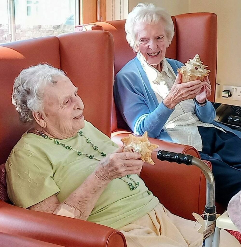 Two elderly women sit in armchairs, smiling and holding star-shaped pastries. Jamaican sunshine streams through the window, filling the room with warmth as they reminisce and enjoy each other's company.