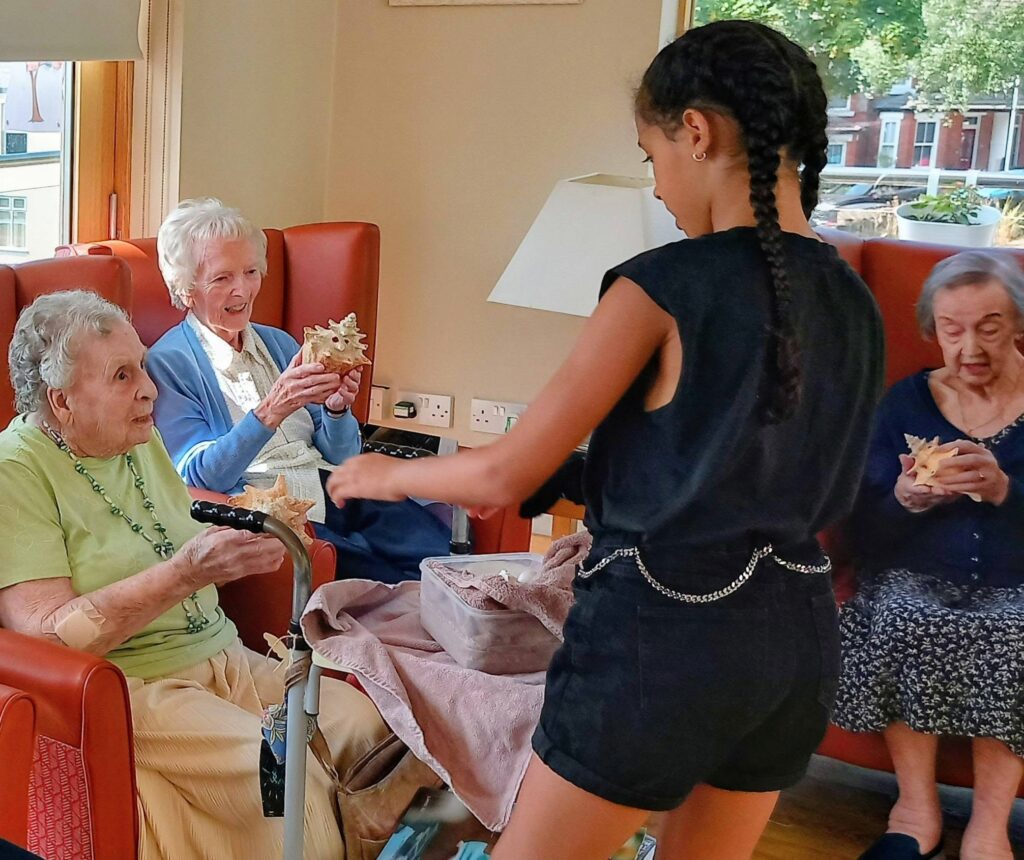 A young girl gives seashells to three elderly women in a care home lounge, where movement is gentle and memories shimmer beneath the warm Jamaican sun shining through large windows overlooking leafy trees.