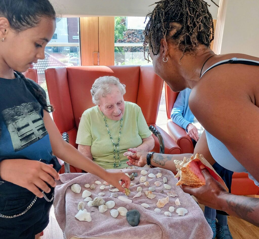 An elderly woman sits smiling in a chair whilst a child and an adult share seashells laid out on a towel-covered table, each shell recalling memories under gentle Jamaican sunshine.
