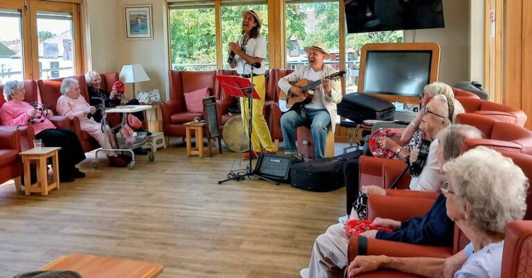 Marko and Andrew perform for a group of elderly people in a lounge, creating a splendid afternoon with music, laughter, and the gentle glow of sunlight streaming through the windows.