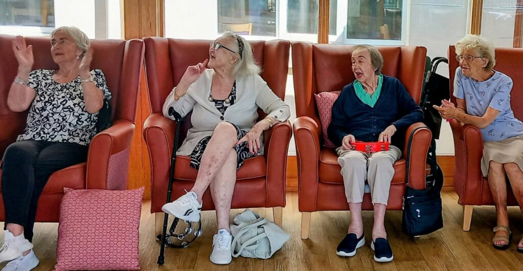 Four elderly women spend a lovely afternoon indoors, seated on brown armchairs. Three chat amiably whilst one holds a red container. Bags and cushions are strewn around them.