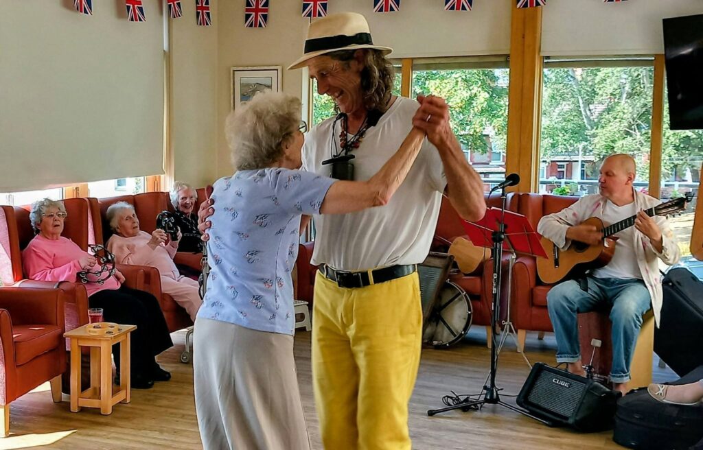 Marko and Andrew dance joyfully in a bright room during a Fabulous Afternoon, as older adults beam from red armchairs beneath festive Union Flag bunting.