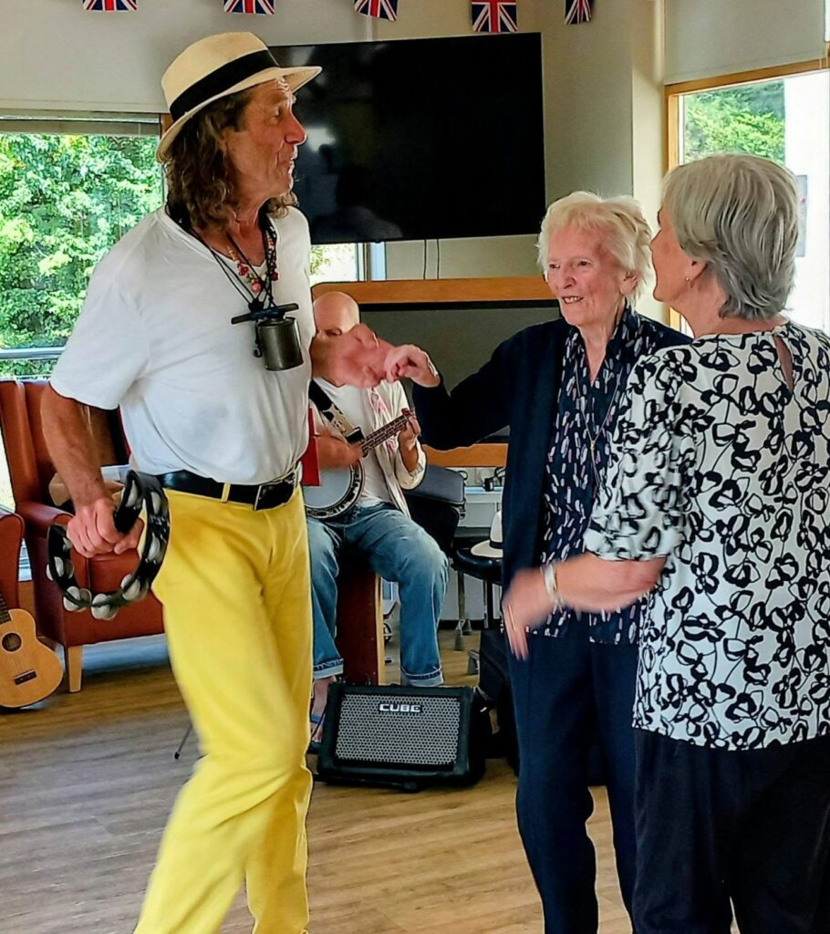 Marko, wearing a hat and yellow trousers, dances with two elderly ladies indoors as a banjo player performs. Union Jack flags hang overhead, setting the scene for a Fabulous Afternoon full of music and happiness.