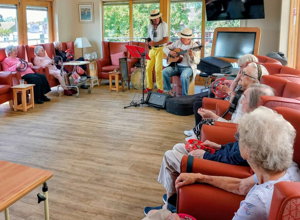 On a sunny afternoon, musicians Andrew and Marko play for elderly people in a communal room; some hold tambourines as sunlight pours through large windows.