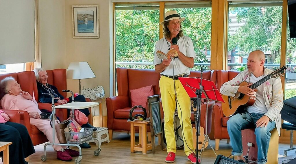 Marko, wearing a hat, reads from a book next to Andrew, the seated guitarist. Several elderly people listen closely in the lounge, enjoying a marvellous afternoon of music and stories.
