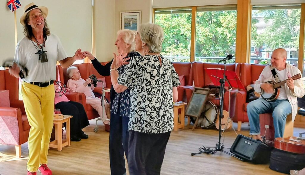 Three elderly women dance joyfully in a bright room on a Fabulous Afternoon, while Marko sings into a microphone, Andrew plays the banjo, and several people watch from their seats in the background.