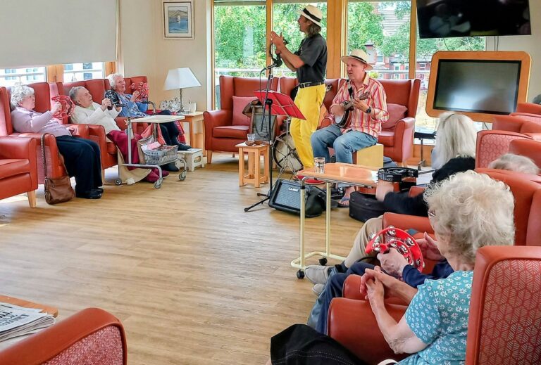 Two musicians perform for a group of elderly people in a well-lit common room. Some audience members clap along to the music, whilst others watch and listen. Musical equipment and furniture can be seen in the background.
