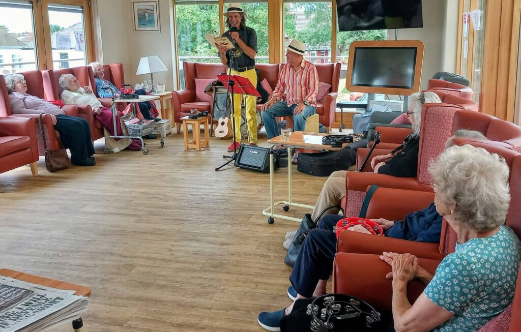 Two musicians fill the bright lounge with music, playing for elderly guests. Some sway gently to the lively tunes, whilst others listen quietly. Musical instruments and equipment are seen around the room.