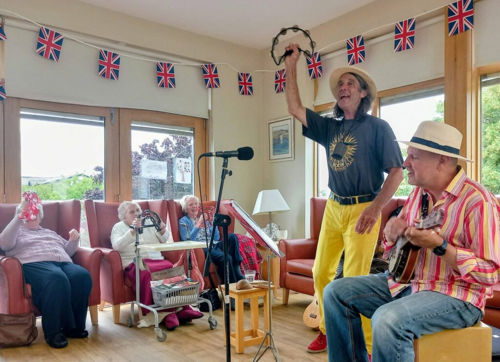 Two musicians entertain elderly people in a lounge adorned with Union Jack flags; one strums a guitar, the other sings and lifts a tambourine. The audience clap, smile, and some begin to dance.