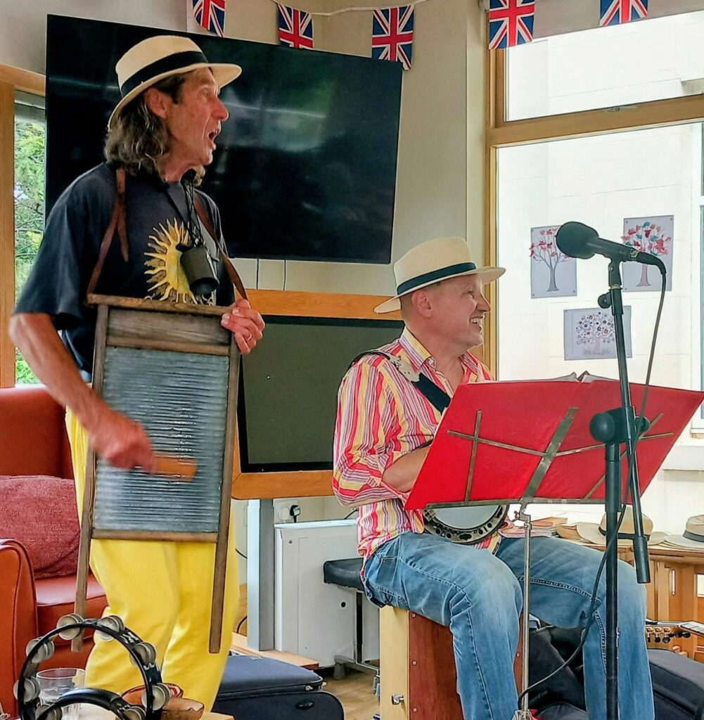 Two musicians perform indoors; one plays a washboard and sings, the other strums a banjo. Both sport hats. Union Jack flags hang above, energising the room with lively music as instruments and microphones rest in the background.
