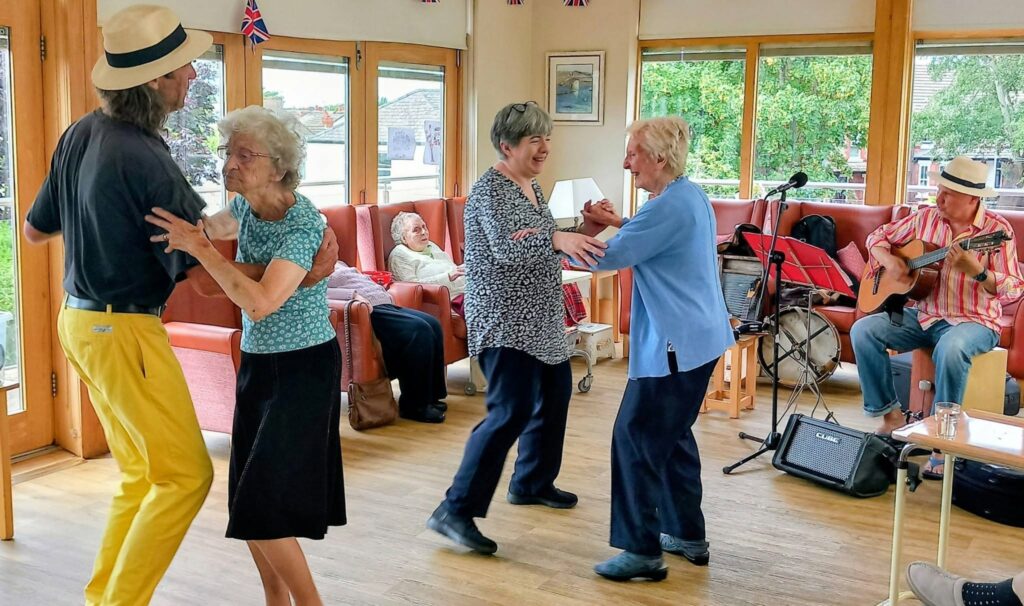 Elderly adults enjoy dancing and music in a bright room with large windows, whilst two musicians play the guitar and sing. Some guests sit back and watch, surrounded by Union Jack flags adding a poetic charm to the scene.
