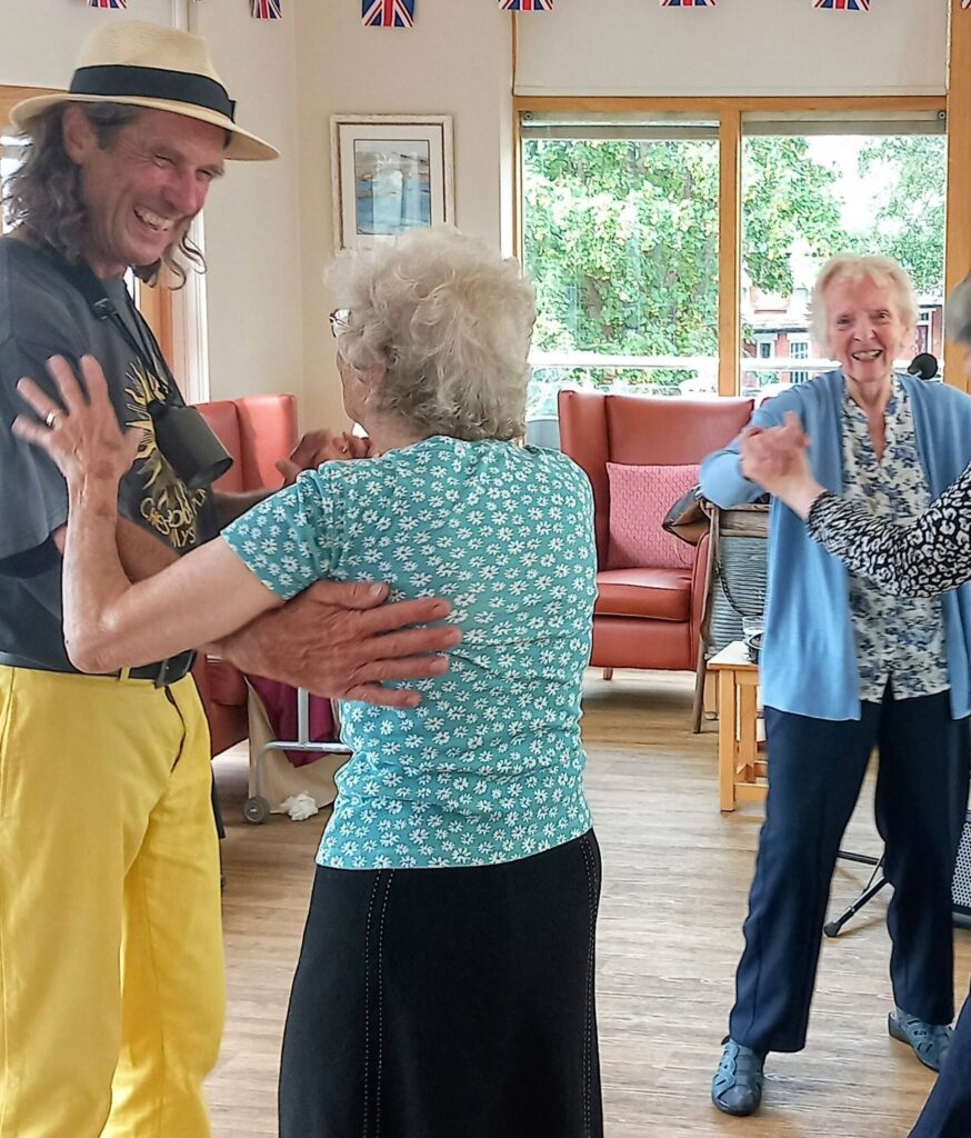 Three older adults dance joyfully together in a bright room with large windows, colourful chairs, and small Union Jack flags hanging on the walls, their movements flowing in time to cheerful music.