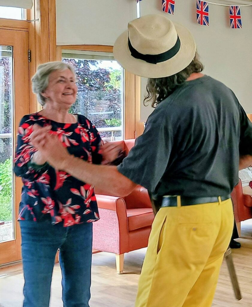 Two people dance together indoors; as music fills the room, the woman beams in a floral blouse and jeans. The man, viewed from behind in a straw hat and yellow trousers, grooves beneath Union Flag bunting.