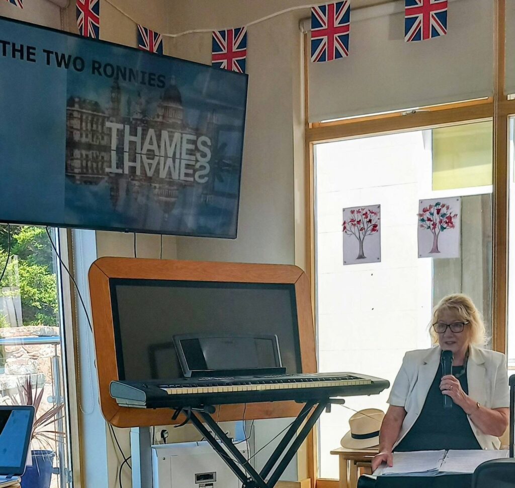 A woman in a white blazer, holding a microphone by a keyboard, reminisces. The Two Ronnies is on the telly near St Paul’s, Union Jacks are draped as Dominga Devitt soaks up the atmosphere.