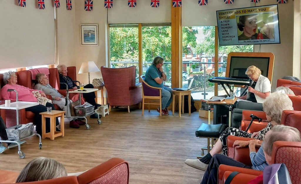 A group of elderly people reminisce in a lounge decorated with British flags, while Dominga Devitt plays the keyboard and sings, lyrics scrolling on the telly. Armchairs and wheelchairs line the cosy room.