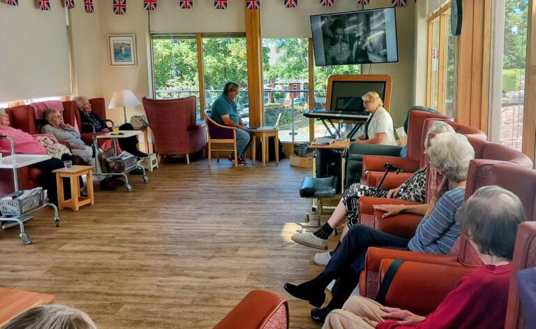 A group of elderly people sit in a communal lounge, enjoying Dominga Devitt’s electric keyboard performance—a real trip down memory lane beneath Union Flag bunting and streaming daylight.