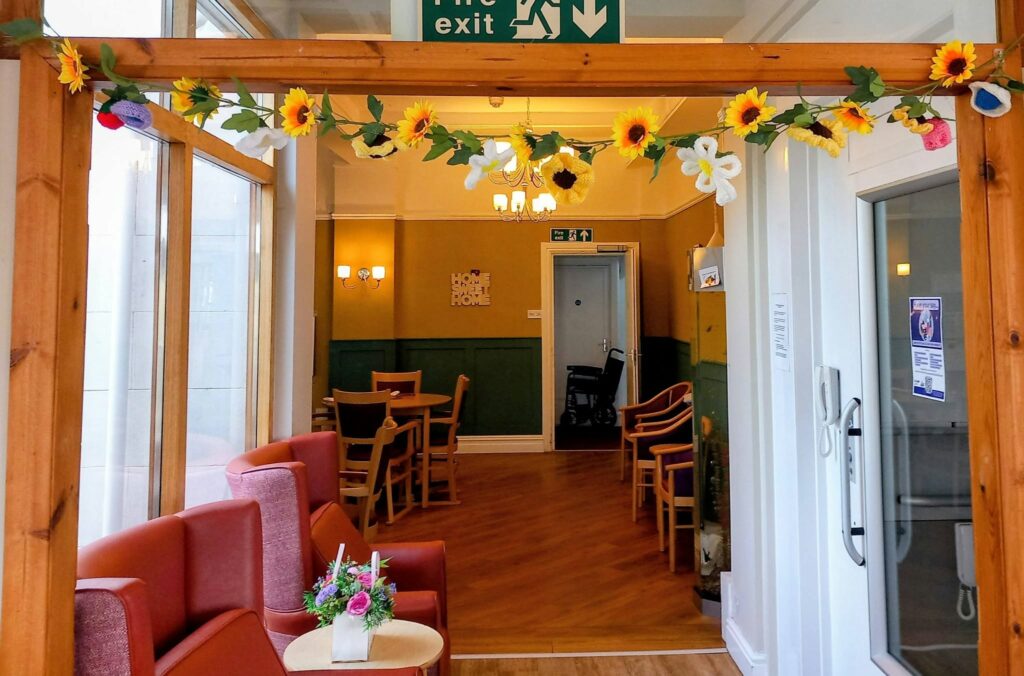 A cosy sitting area with red chairs, wooden tables, and a floral garland above the entrance invites you to knit and natter. Lamps line the walls near a Home Sweet Home sign, with an emergency exit sign visible.