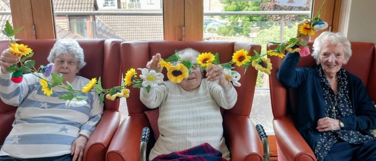 Three elderly women sit in armchairs, holding up a vibrant floral garland adorned with artificial flowers and leaves—a real team effort. Large windows and nearby houses are visible in the background.