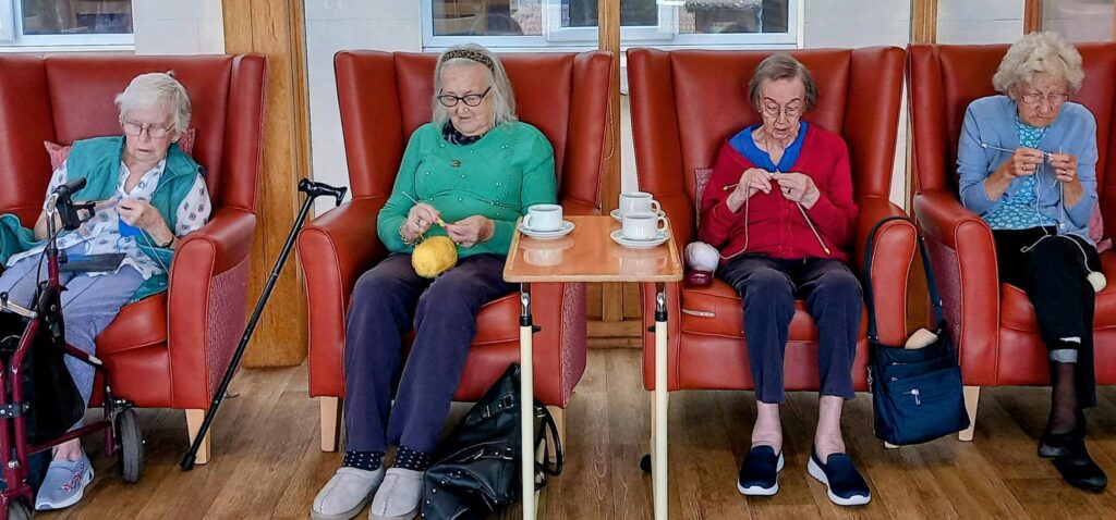 Four elderly women knit together on red chairs, their teamwork clear as they chat. A wooden table with teacups stands between them, whilst walking frames and bags sit nearby in sunlight streaming from large windows.