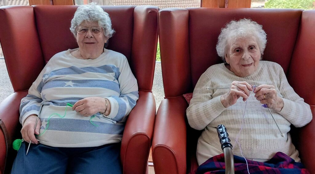 Two elderly women sit in large red armchairs, each with knitting needles and wool, pausing their chat to offer a slight smile at the camera.