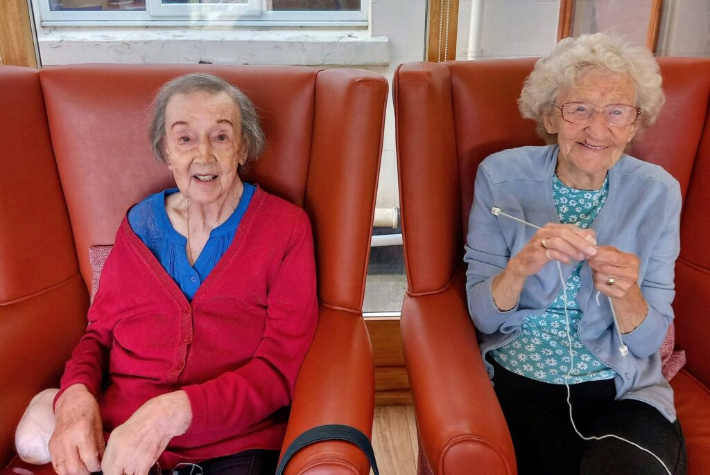 Two elderly women sit side by side in brown armchairs. One, wearing a red cardigan, smiles at the camera; the other, dressed in blue, smiles and holds knitting needles with wool—a real team effort as they knit together.