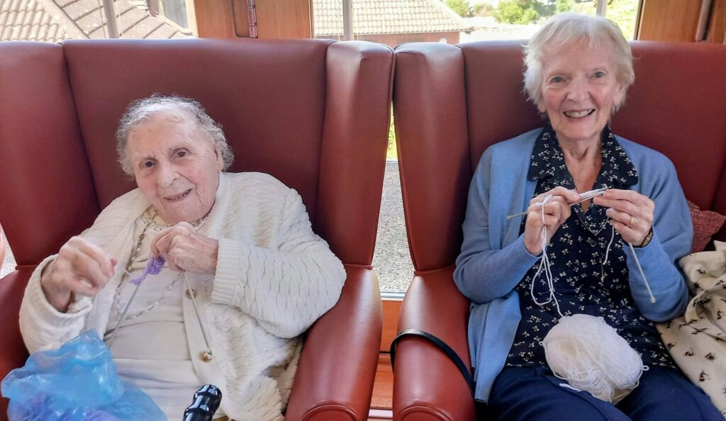 Two elderly women sit together on red chairs, smiling as they knit and have a natter, holding wool and needles. Gentle natural light pours in through the window behind them.