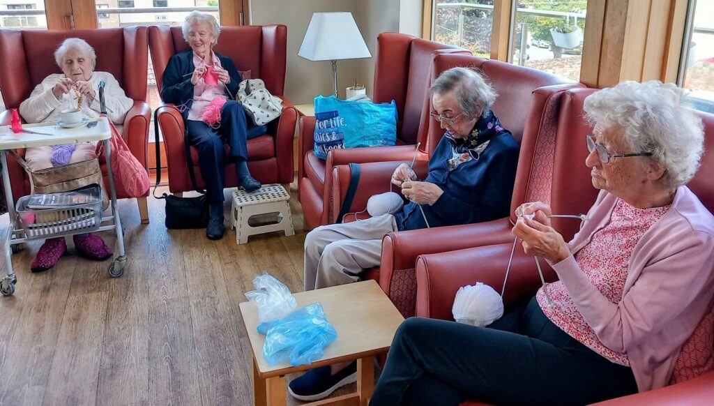 Four elderly women sit knitting and having a natter in armchairs, surrounded by white wool and a blue bag. A walking frame is nearby, as a floral garland adds colour to the serene, well-lit room.