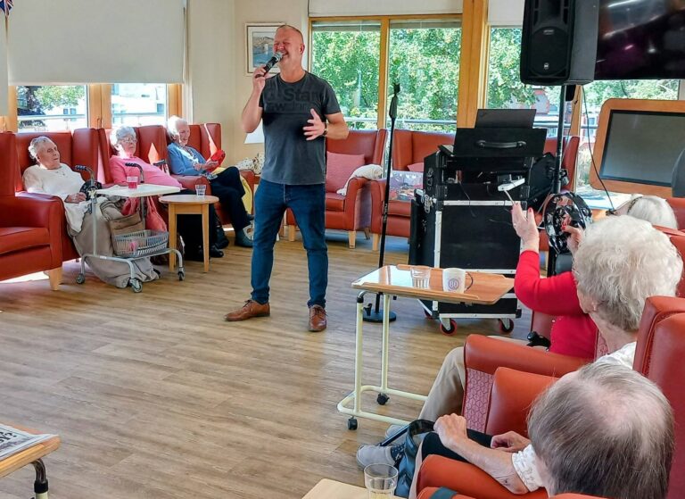 Mark Birch sings into a microphone at Lear House, entertaining elderly people seated in armchairs. Audio equipment sits close by as some clap and enjoy the lively performance.