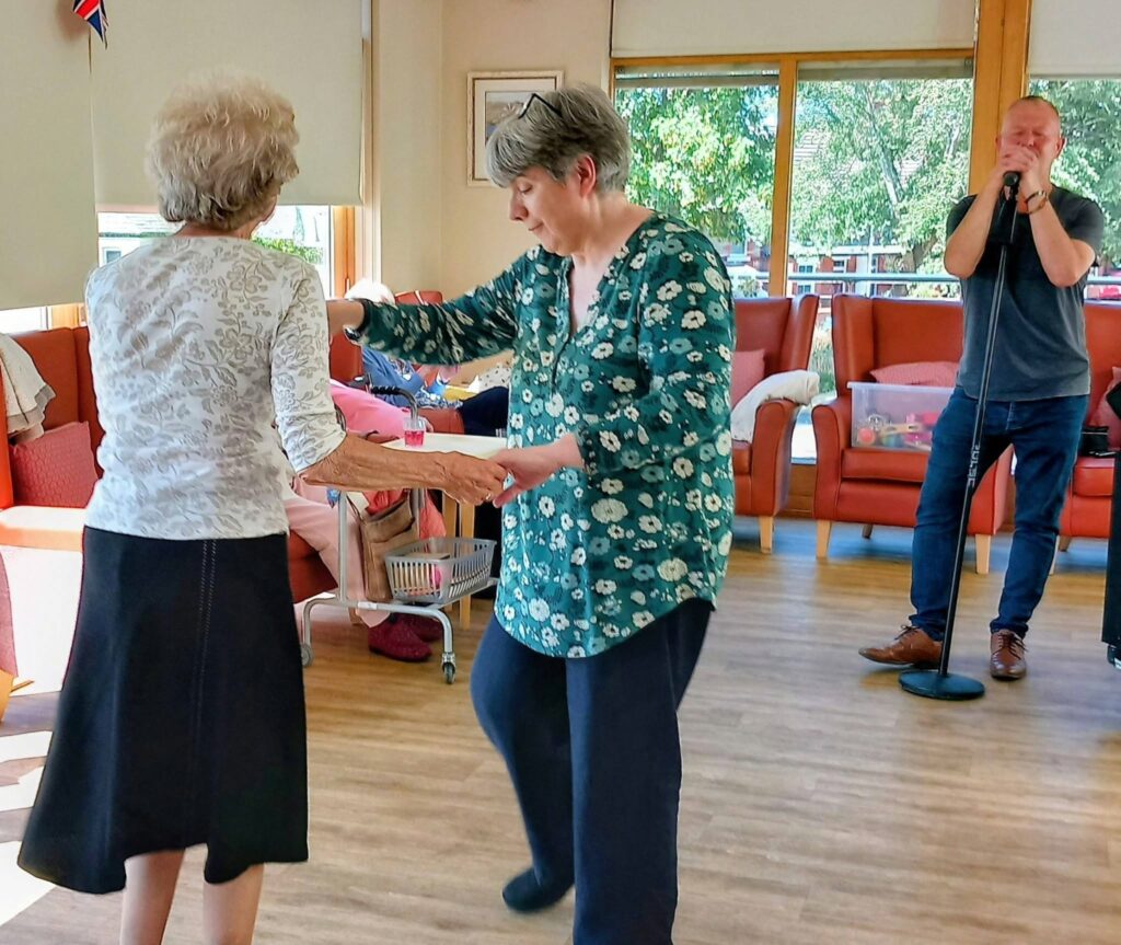 Two women dance in the sunlit Lear House as a man, possibly Mark Birch, sings into a microphone. Red chairs and large windows surround them, with music and natural light filling the atmosphere.