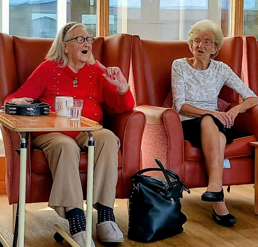 Two elderly women sit in red armchairs by the window at Lear House. One gestures as she speaks, with music playing softly in the background; a tray and black bag are nearby.