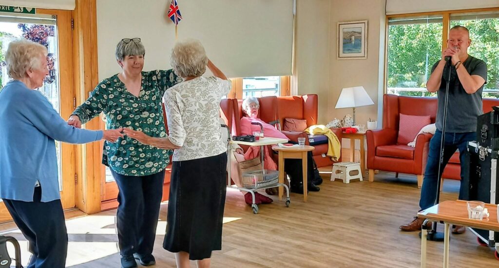 At Lear House, three elderly women dance to music while Mark Birch plays the harmonica. Another woman watches from a chair in the cosy, sunlit room decorated with a small UK flag.