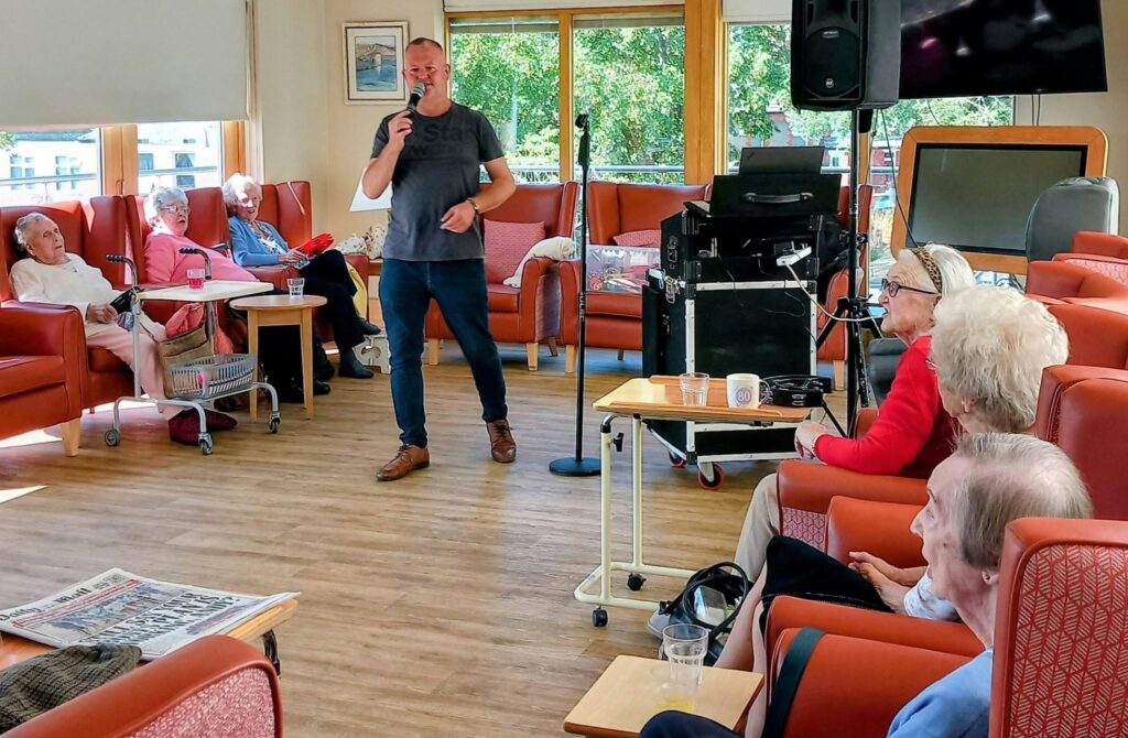 Mark Birch sings into a microphone at Lear House, as elderly residents enjoy the music from red chairs. Sunlight streams through large windows and audio equipment is positioned nearby.
