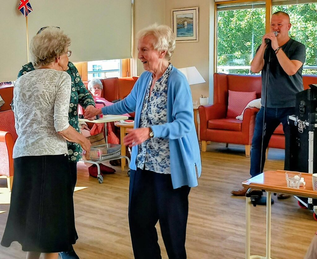 Two elderly women dance together at Lear House while Mark Birch plays live music. In the bright room, people sit in armchairs near large windows, soaking up the lively atmosphere.