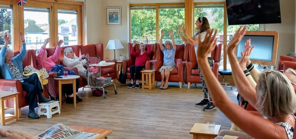 A group of elderly people sit in a circle in a bright room, lifting their arms as they follow a woman leading a gentle exercise class with music and movement.