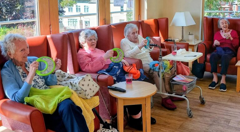At Lear House, four elderly women sit comfortably in armchairs; three take gentle exercise with green rings whilst one drinks from a cup. Sunlight streams through large windows as music and movement promote well-being.
