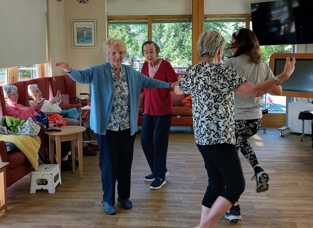 Several elderly women and a younger woman take part in gentle exercise, dancing together in a bright communal lounge, while other pensioners relax on sofas by large windows.