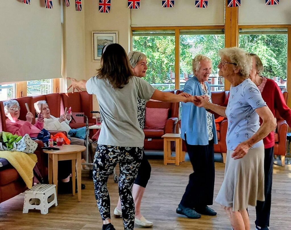 Several elderly women enjoy music and movement in a brightly lit room decorated with Union Jack bunting, whilst others sit on sofas, watching the lively scene unfold.