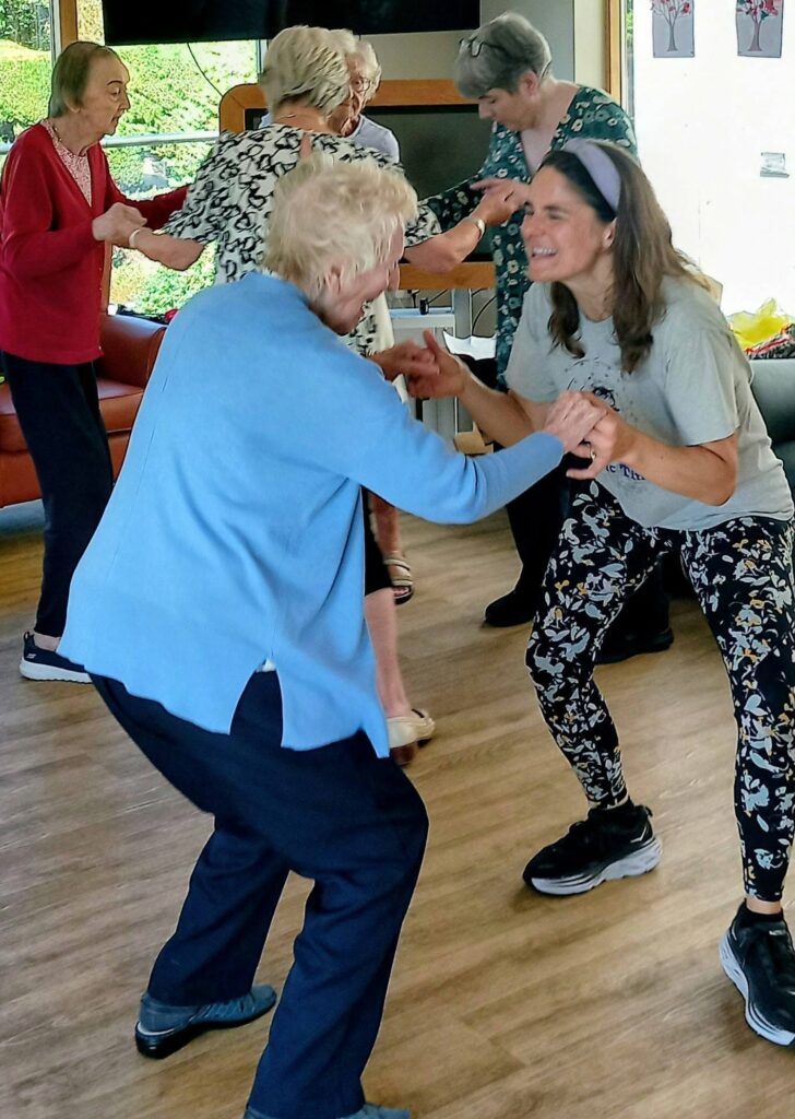 A group of older adults and a younger woman hold hands and dance together, enjoying music and movement in a bright room with wooden flooring and comfortable seating.
