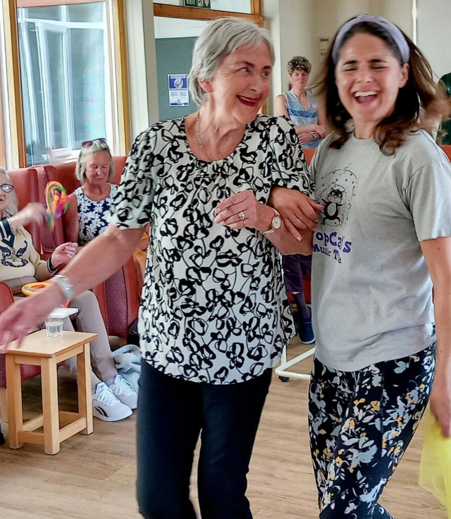 Two women smile and walk arm-in-arm at Lear House, where others enjoy gentle exercise in a bright, cheerful room. The relaxed atmosphere is filled with socialising, music and movement.