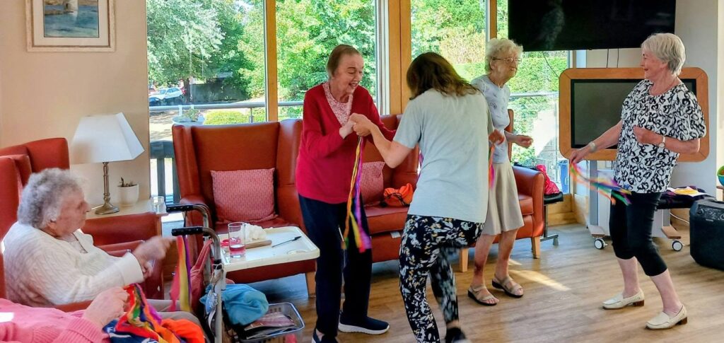 Five elderly women in a brightly lit room enjoy gentle exercise with colourful ribbon sticks; some stand and dance along to music and movement, whilst one woman sits and watches.
