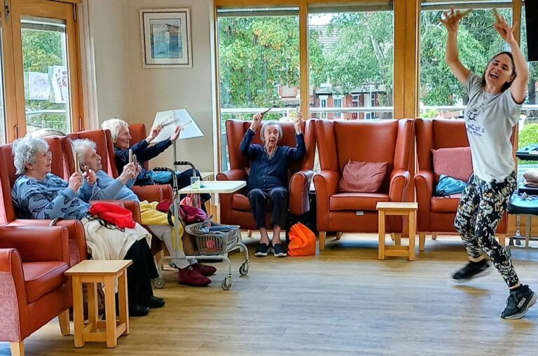 A group of elderly people sit in armchairs, clapping along to music and movement as a younger woman leads an exercise or dance session in a bright, communal room with large windows.