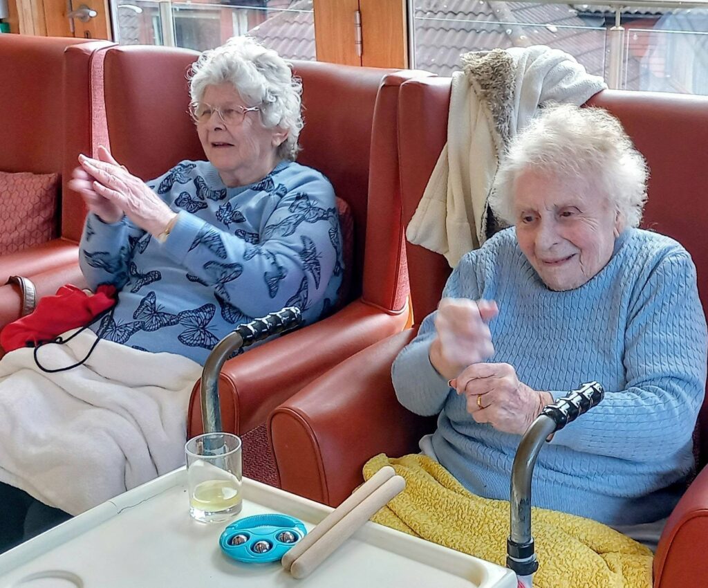 Two elderly women sit together in armchairs, hand jiving and clapping along to music and movement activities. A small table between them holds a glass of water, toy bells, and wooden sticks.