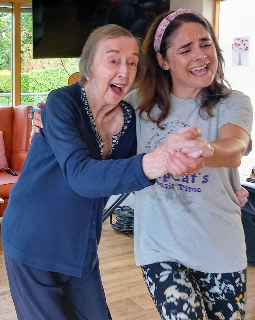 An elderly woman and a younger woman smile and dance indoors, hand jiving cheerfully. The younger woman sports a headband and T-shirt, enjoying music and movement in this touching moment.