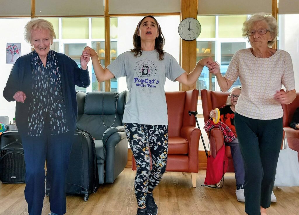 Three women waltz, holding hands and dancing in a brightly lit Lear House room; two are older, one younger wearing a PopCats Music Time T-shirt. A clock and people seated are visible in the background.